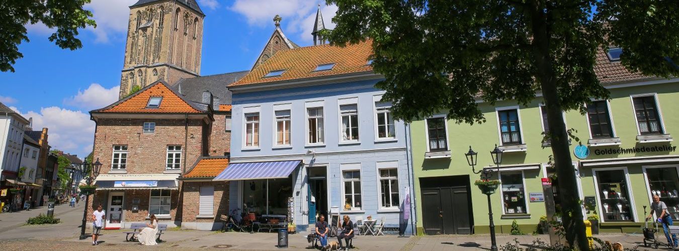 Viersen (Suchteln): Blick über den Marktplatz auf mittelalterliche Häuser und Geschäfte mit Kirchturm im Hintergrund.