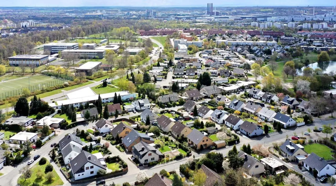 Luftaufnahme einer Wohnsiedlung am Stadtrand von Wolfsburg in Deutschland mit einer Schule im Hintergrund und Fabriken am Horizont.