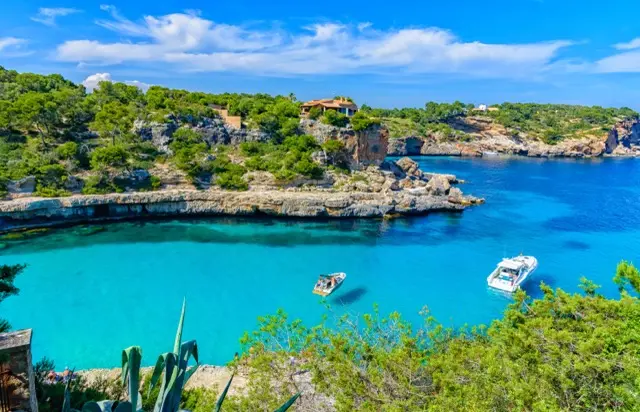 Türkisfarbene Bucht auf Mallorca mit zwei Booten, Felsküste, Pinien und einer Finca auf der Landzunge