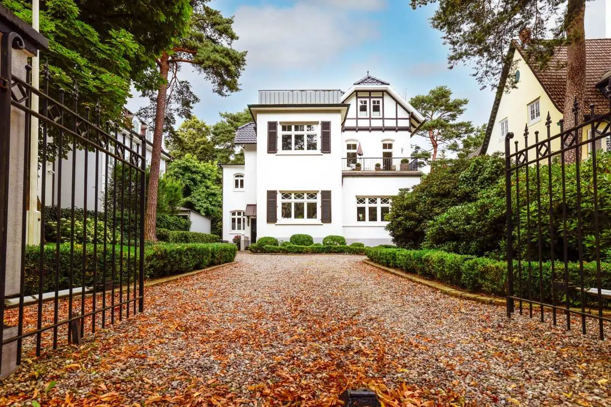 Weiße Jugendstil-Villa mit Fachwerkgiebel und Klappläden hinter schmiedeeisernem Tor, gepflegter Kiesauffahrt und Buchsbaumhecken im Herbst