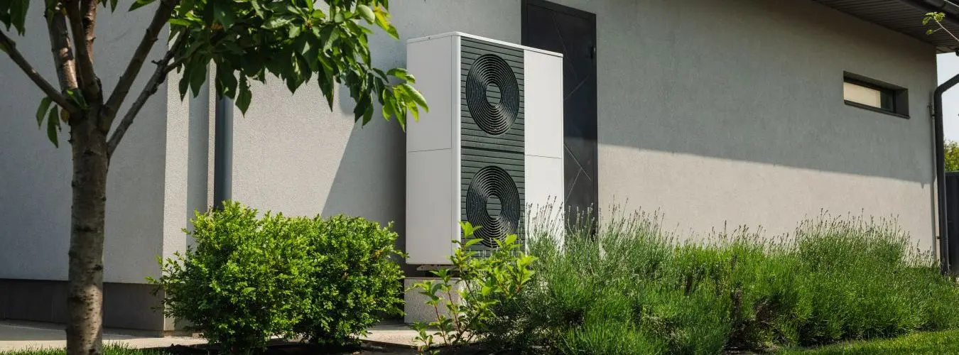 Moderne Luft-Wärmepumpe an der Außenwand eines grauen Einfamilienhauses mit angelegtem Garten
