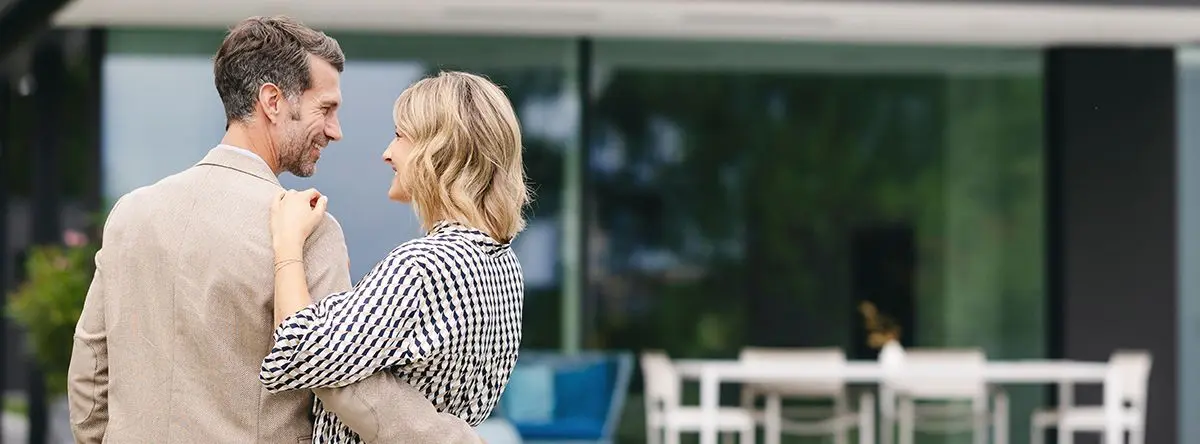 Glückliches Paar umarmt sich vor einem modernen Haus mit Terrasse und Garten – Symbolbild für die erfüllte Finanzierung des Eigenheims
