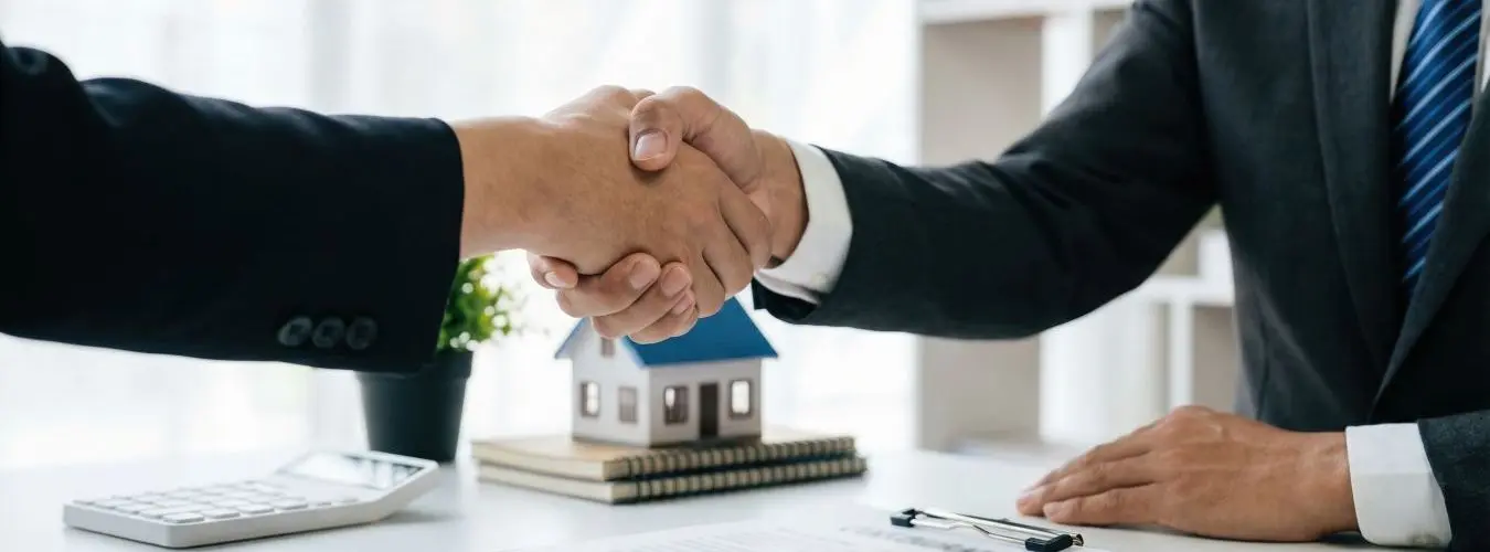 Handschlag zwischen Immobilienmakler und Kunde nach erfolgreichem Vertragsabschluss, im Hintergrund ein Hausmodell auf Vertragsunterlagen